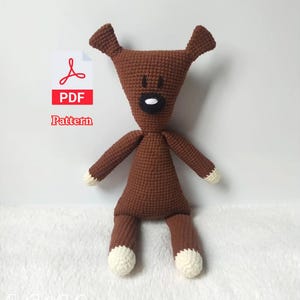 Può includere: Un cane all'uncinetto marrone fatto a mano con dettagli bianchi sulle zampe e sulle punte delle braccia. Il cane ha un naso nero e una bocca bianca. L'immagine include un'icona del modello PDF.