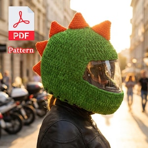 Könnte beinhalten: Ein grüner, gehäkelter Motorradhelmbezug mit orangefarbenen Stacheln, der einem Dinosaurier ähnelt. Der Helm hat ein klares Visier. Das Bild enthält den Text "PDF Pattern". Die Person trägt eine schwarze Lederjacke.