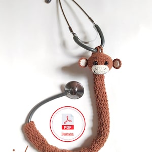 Puede incluir: Un estetoscopio con una funda de mono de ganchillo marrón. El mono tiene detalles blancos en la cara y las orejas. El estetoscopio es plateado y gris. La imagen también incluye un logotipo de patrón PDF y la palabra "icha".