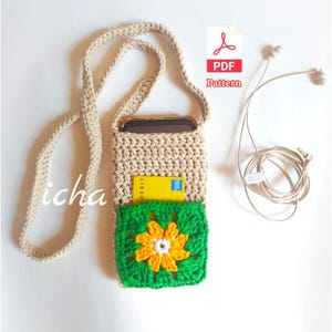 May include: A beige crochet phone crossbody bag with a green and yellow flower design on the front pocket. A yellow debit card is visible in the pocket. The bag has a long strap and is next to a pair of beige earbuds.