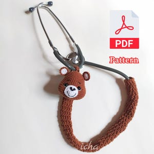 May include: A gray metal stethoscope featuring a brown crocheted bear cover on the tubing. The bear head has small round ears, black eyes, and a white muzzle with a black nose and mouth. A PDF Pattern icon is visible in the upper right corner.