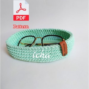 May include: A light green crocheted basket holding eyeglasses. The basket has a rounded shape and is made of a textured yarn. The eyeglasses have black frames and gold-colored arms. A small leather tag is attached to the basket. The word "icha" is written on the basket.