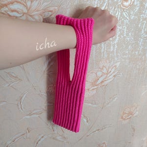Könnte beinhalten: Ein leuchtend pinkfarbener Handgelenkwärmer aus Rippstrick mit fingerlosem Design. Der Text "icha" ist auf dem Arm zu sehen. Der Handgelenkwärmer besteht aus einem weichen, strukturierten Material.
