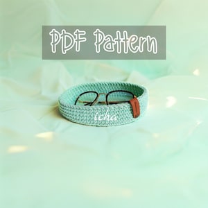 May include: A crocheted, light green basket holds a pair of eyeglasses with a brown leather tag. The text "PDF Pattern" is displayed above the basket. The word "icha" is written on the basket.