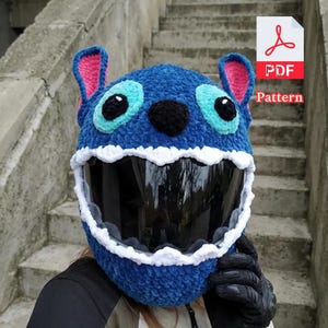 May include: A blue crocheted motorcycle helmet cover, designed to resemble a cartoon character. It has large, turquoise eyes, a black nose, and white teeth around the visor. The ears are blue with pink interiors. Includes a PDF pattern icon.