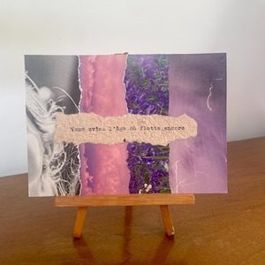 Può includere: Un'opera d'arte a tecnica mista su un piccolo cavalletto di legno. L'opera presenta strisce di carta strappate nei toni del rosa, viola e grigio, con una sezione di fiori viola. Il testo in francese recita: "Vous aviez l'âge où flotte encore."
