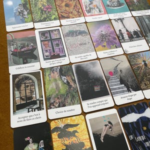 May include: A collection of tarot cards with various illustrations and text. The cards feature images of people, nature, and abstract designs. The text on the cards is in French. The cards are arranged on a brown surface.