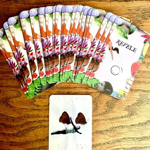 May include: A deck of tarot cards fanned out on a wooden surface. The cards feature colorful illustrations of figures, plants, and animals. One card shows a person with butterfly wings. The word "REVELE" is visible on one card, and "Surmonter nos peurs" on another.