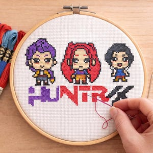 Puede incluir: Bastidor de bordado de punto de cruz con tres personajes de dibujos animados y la palabra "HUNTER" en morado y rojo. Hilos de bordar coloridos y tijeras doradas.
