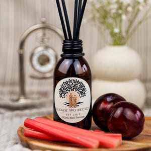 May include: A brown glass bottle with a black label and a tree design. The label reads "Flicker Apothecary Est 2025 100ml Reed Diffuser". The bottle is surrounded by red rhubarb stalks and two dark red plums on a wooden surface.