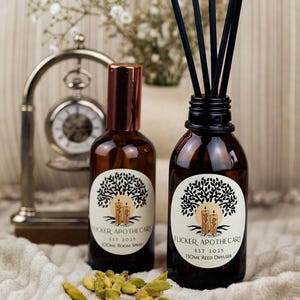 Peut inclure: Deux flacons en verre ambré avec des étiquettes brunes et des bouchons noirs. Les étiquettes représentent un arbre avec un soleil et des bougies. Le texte sur les étiquettes indique "Flicker Apothecary Est 2023" et "100ml Room Spray" sur un flacon et "Flicker Apothecary Est 2023" et "130ml Reed Diffuser" sur l'autre flacon. Les flacons sont posés sur une surface en tissu blanc avec des gousses de cardamome vertes éparpillées autour.
