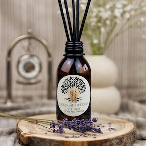 May include: A brown glass bottle with a black label that reads "Flicker Apothecary Est 2025 120ml Reed Diffuser". The bottle is filled with a clear liquid and has black reeds sticking out of the top. The bottle is sitting on a wooden surface with lavender flowers.