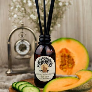 May include: A brown glass reed diffuser bottle with black reeds, a white label with a tree and candle design, and the text "FLICKER APOTHECARY EST 2025 130ML REED DIFFUSER". Sliced cantaloupe and cucumber are in the foreground. A silver clock and white flowers are in the background.