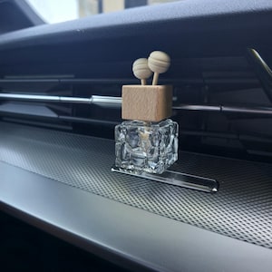 May include: A car air freshener featuring a clear, geometric glass bottle, a wooden cube, and two spherical wooden toppers. The freshener is placed on a silver dashboard with a perforated pattern.