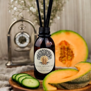 May include: A brown glass bottle with a black label that reads "Flicker Apothecary Est 2023 130ml Reed Diffuser". The bottle is surrounded by cantaloupe slices and cucumber slices on a wooden surface.