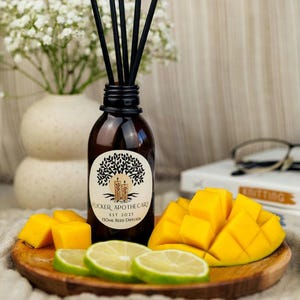 May include: A brown glass bottle with a black label that reads "Flicker Apothecary Est 2023 Floral Reed Diffuser". The bottle is filled with a clear liquid and has black reeds sticking out of the top. The bottle is sitting on a wooden surface with sliced mango and lime wedges.