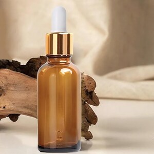 May include: Amber glass dropper bottle with a gold collar and white dropper top. The bottle is empty and sits on a white surface with a neutral background. The bottle is likely for essential oils or serums.