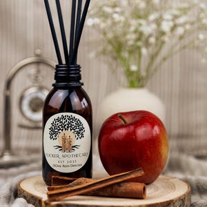 Peut inclure: Un diffuseur de roseaux en verre brun avec des roseaux noirs, une pomme rouge et des bâtons de cannelle sur une tranche de bois. La bouteille porte l'inscription "Flicker Apothecary EST 2025 130ml Reed Diffuser".