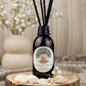 May include: A brown glass bottle with a white label featuring a tree and the text "Flicker Apothecary Est 2023 130ml Reed Diffuser". The bottle is filled with a clear liquid and has black reeds sticking out of the top. The bottle is sitting on a wooden surface with white flowers and seashells.