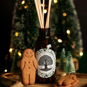 May include: A brown glass reed diffuser bottle with wooden reeds, a gingerbread man biscuit, and a stack of gingerbread biscuits. The bottle has a white label with a tree design. The background includes a Christmas tree and lights.