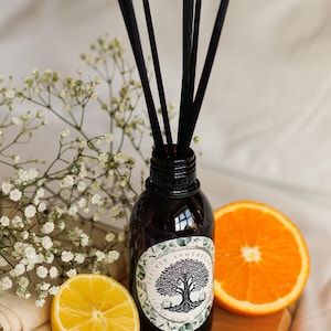 May include: A dark brown glass diffuser bottle with black reeds, a label with a tree design, and a half orange and lemon on a wooden board. The bottle is surrounded by white flowers.