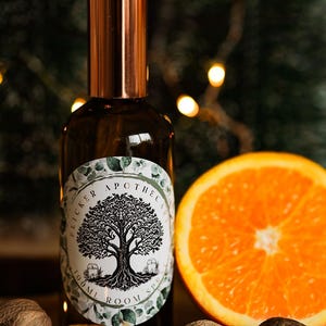 Peut inclure: Un flacon de spray d'ambiance en verre brun avec un bouchon en cuivre et une étiquette avec un motif d'arbre. Une tranche d'orange, des bâtons de cannelle et de la noix de muscade sont disposés à proximité. L'étiquette indique "Flicker Apothecary 100ml Room Spray."