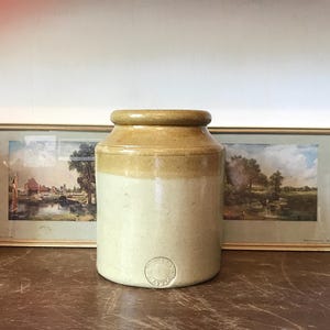 May include: A vintage stoneware jar with a light brown glaze and a white base. The jar has a circular design on the front. The jar is sitting on a wooden surface with two vintage glass bottles on either side.