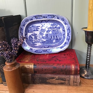 May include: A blue and white Willow pattern serving dish sits atop a stack of antique books. A small brown vase holds dried lavender, and a silver candlestick with a yellow candle is to the right.