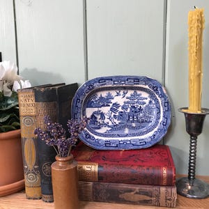 Puede incluir: Un plato ovalado de cerámica azul y blanco con un diseño de paisaje se encuentra sobre una pila de libros antiguos. A la derecha hay una vela alta amarilla en un candelabro plateado. Un pequeño jarrón marrón contiene lavanda.