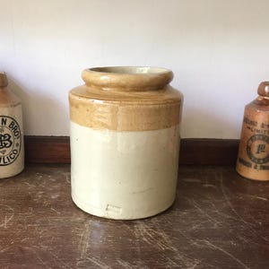 May include: A vintage stoneware jar with a tan rim and a cream body. Two smaller bottles with labels and a red book are either side of the jar. The items are on a wooden surface.