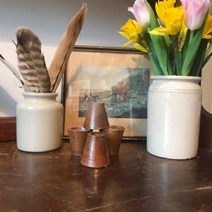 Puede incluir: Una estantería de madera con un jarrón de cerámica blanca lleno de narcisos y tulipanes amarillos y rosas. Hay dos tarros de cerámica blanca en la estantería, uno con plumas y el otro vacío. Hay cuatro pequeñas tazas de madera en la estantería delante de un cuadro enmarcado.