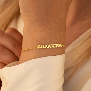 May include: A delicate gold bracelet spelling out the name "ALEXANDRA" in capital letters. The bracelet is attached to a fine chain and is worn on a wrist, against a white shirt.