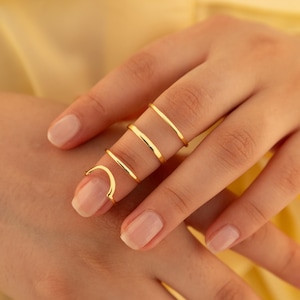 May include: Close-up of a hand wearing four gold rings. The rings are thin and minimalist in design, with one ring curving around the fingernail. The rings are a bright gold color, and the hand has a neutral nail polish.