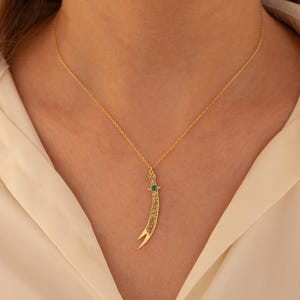 May include: A gold-colored necklace featuring a pendant shaped like a curved sword. The pendant has an emerald-colored stone and intricate detailing. The necklace is displayed against a white background.