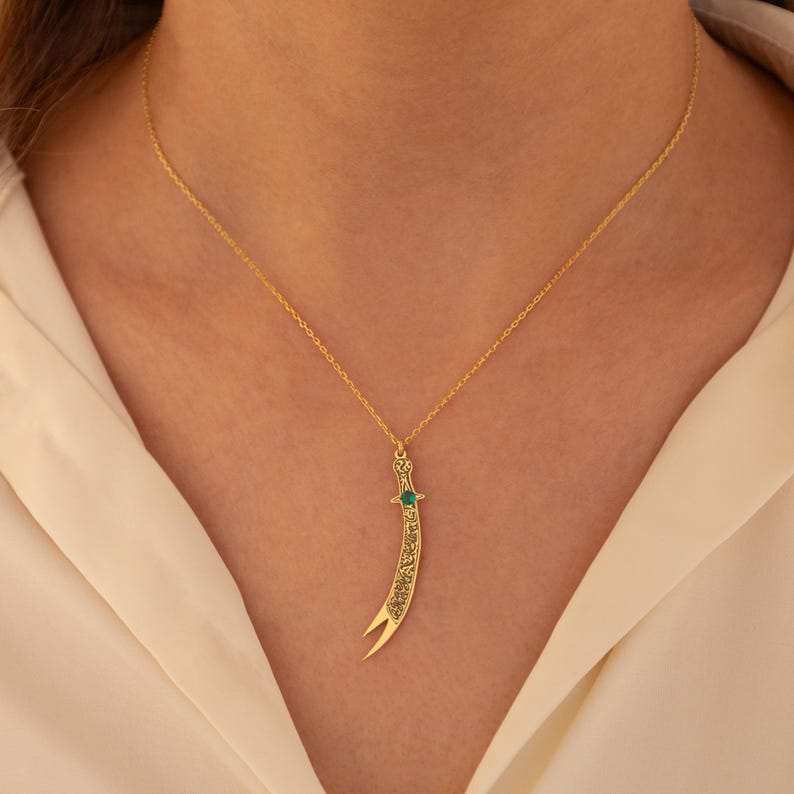 May include: A gold-colored necklace featuring a pendant shaped like a curved sword. The pendant has an emerald-colored stone and intricate detailing. The necklace is displayed against a white background.