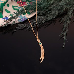 May include: A gold-colored pendant necklace featuring a curved dagger design. The dagger has intricate etched details and a small green gemstone. The necklace hangs on a delicate chain, set against a dark background.