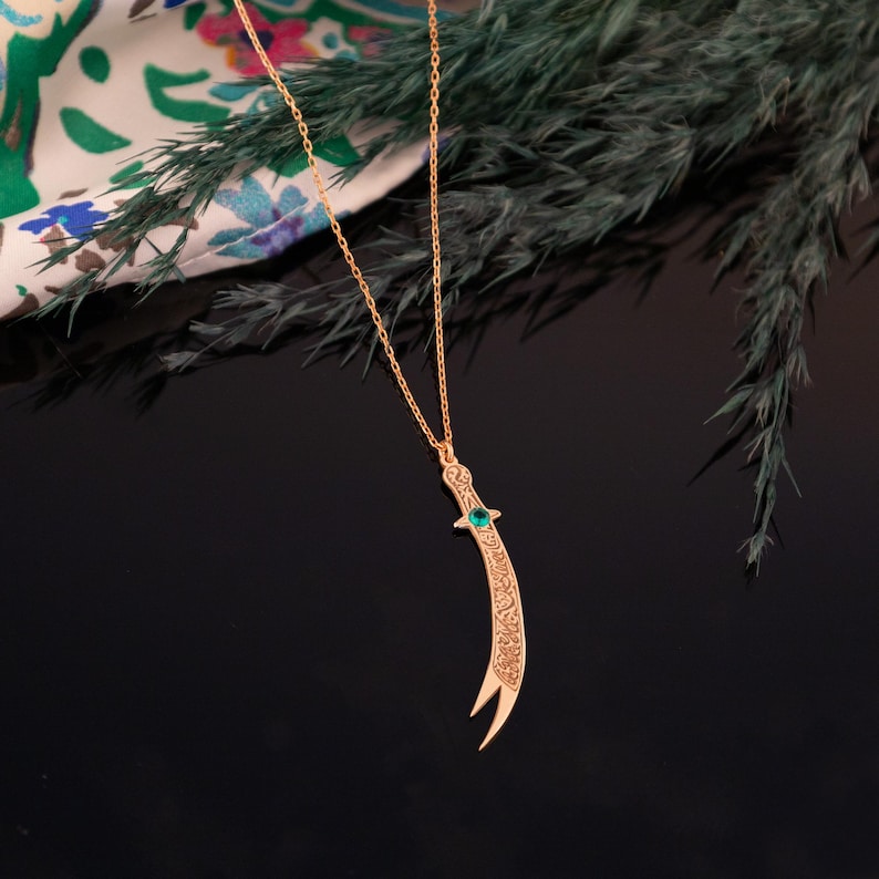 May include: A gold-colored pendant necklace featuring a curved dagger design. The dagger has intricate etched details and a small green gemstone. The necklace hangs on a delicate chain, set against a dark background.