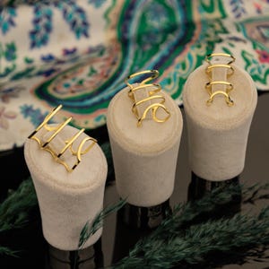 May include: Three gold-colored rings on display. Each ring has a unique design, including a spiral, a series of connected arches, and a geometric shape. The rings are presented on small, off-white pedestals against a patterned fabric background.