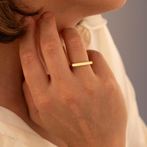 May include: A gold-colored ring with a rectangular bar design. The ring is worn on a finger, showcasing its simple, modern aesthetic. The jewelry piece is a minimalist accessory.