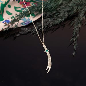 May include: Silver-toned pendant necklace featuring a curved dagger design. The dagger has intricate detailing and a green gemstone accent. The necklace hangs on a delicate chain, set against a dark background with greenery and a patterned fabric.