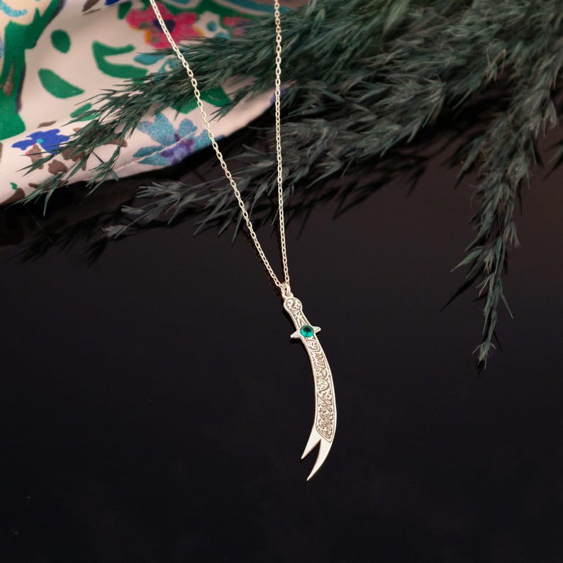 May include: Silver-toned pendant necklace featuring a curved dagger design. The dagger has intricate detailing and a green gemstone accent. The necklace hangs on a delicate chain, set against a dark background with greenery and a patterned fabric.