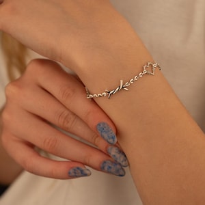 May include: Silver-toned bracelet featuring a barbed wire design with a heart-shaped charm. The bracelet is worn on a wrist with blue and white nail polish.