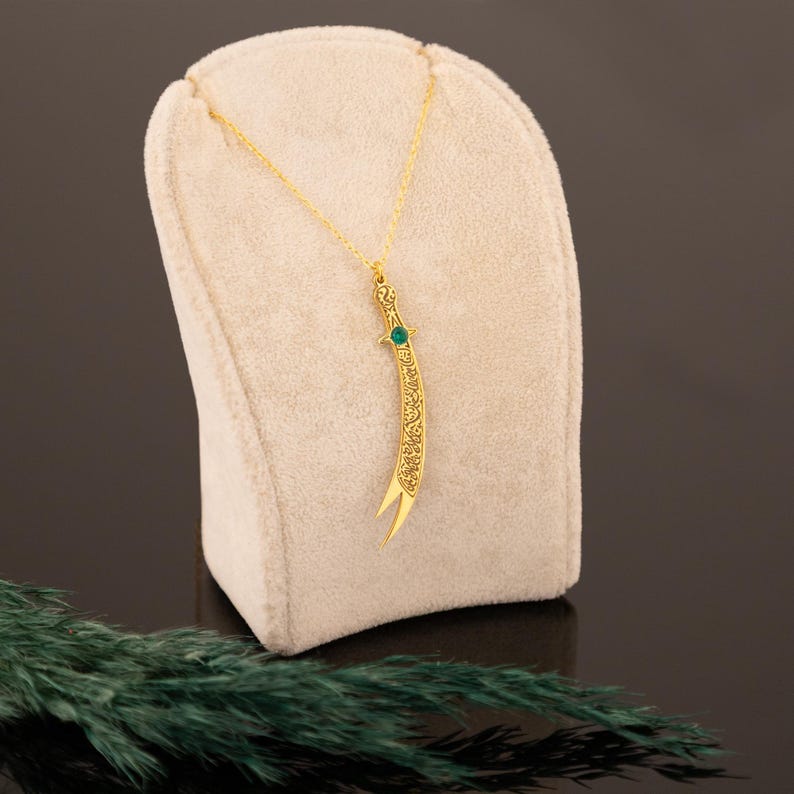 May include: A gold-colored pendant necklace featuring a curved sword design. The sword has intricate detailing and a small green gemstone. The necklace is displayed on a beige stand against a dark background.