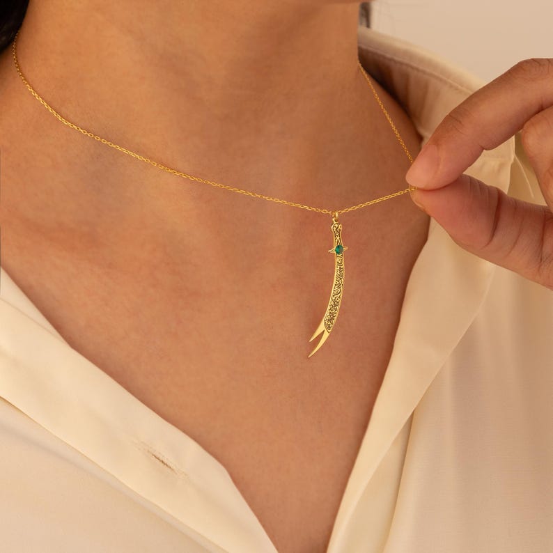 May include: A gold-colored necklace featuring a pendant shaped like a curved sword. The pendant has intricate etched designs and a small green gemstone. The necklace is worn against a white shirt.