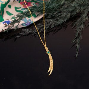 May include: A gold-colored pendant necklace featuring a curved dagger design. The dagger has intricate detailing and a small green gemstone. The necklace hangs on a delicate gold chain, set against a dark background.