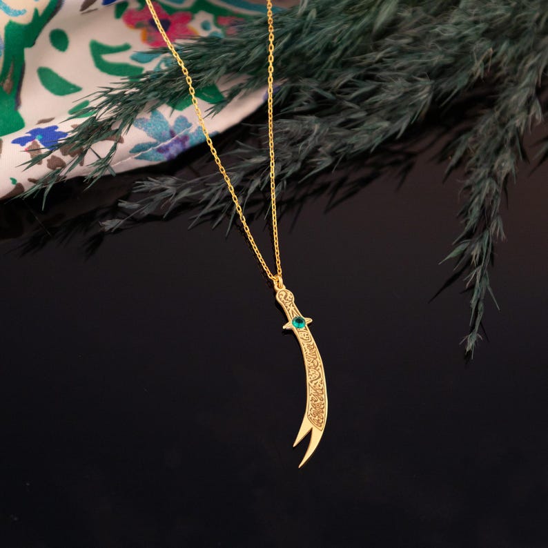 May include: A gold-colored pendant necklace featuring a curved dagger design. The dagger has intricate detailing and a small green gemstone. The necklace hangs on a delicate gold chain, set against a dark background.