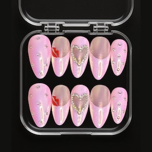 May include: A set of ten pink, iridescent, almond-shaped press-on nails in a clear plastic case. Some nails feature a rhinestone heart, a red lip design, or scattered rhinestones. The nails are designed for a glamorous look.