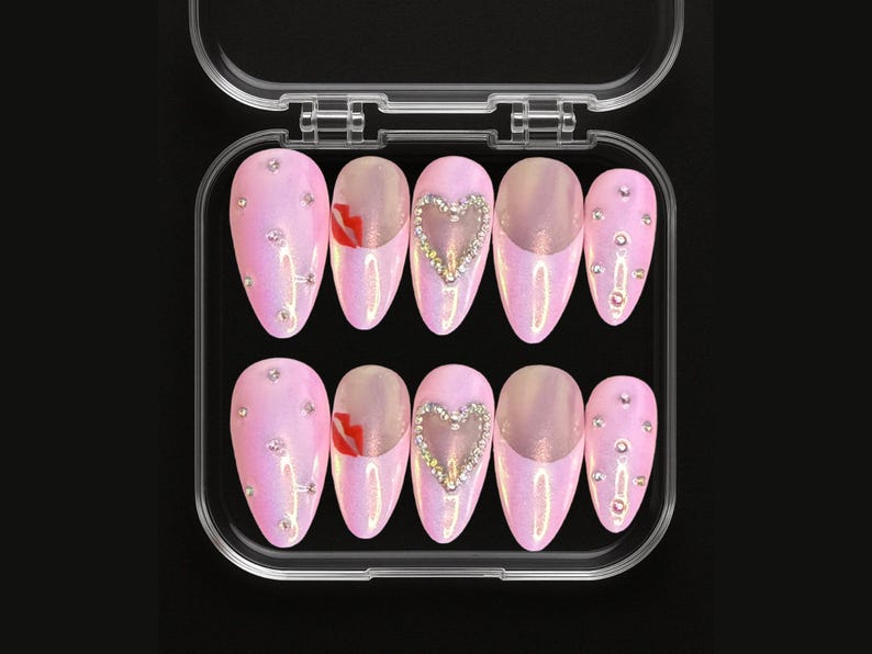 May include: A set of ten pink, iridescent, almond-shaped press-on nails in a clear plastic case. Some nails feature a rhinestone heart, a red lip design, or scattered rhinestones. The nails are designed for a glamorous look.