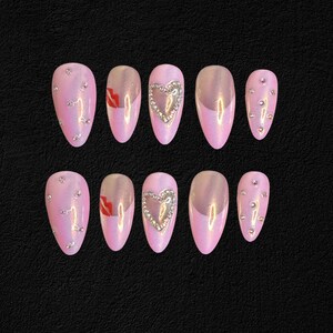 May include: Set of ten pink press-on nails. Some nails feature silver rhinestones, a silver heart design, or a red lip design. The nails have an iridescent finish and are arranged on a black surface.