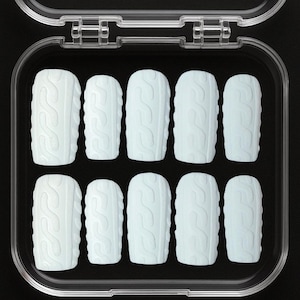 May include: A set of ten light blue press-on nails with a cable knit design, arranged in a clear plastic case. The nails are rectangular with rounded edges, and the case is open, revealing the full set against a black background.
